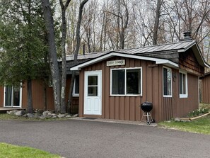 Une petite cabane à un seul étage, avec une porte et des fenêtres blanches, une cheminée et un barbecue à l’extérieur.