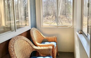 A corner room with two wicker chairs, a window with a view of a forest, and a door.