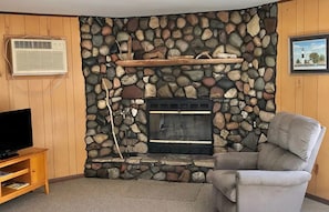 A stone fireplace with a mantel, a television on a wooden stand, and a cushioned armchair.