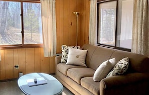 A room with a wooden paneled wall, a beige sofa with patterned cushions, a small round table with a remote control, and a tall floor lamp.