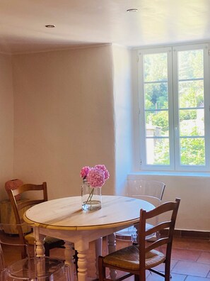 Et spisebord med stole, en vase med blomster og et vindue med udsigt til grøn vegetation.