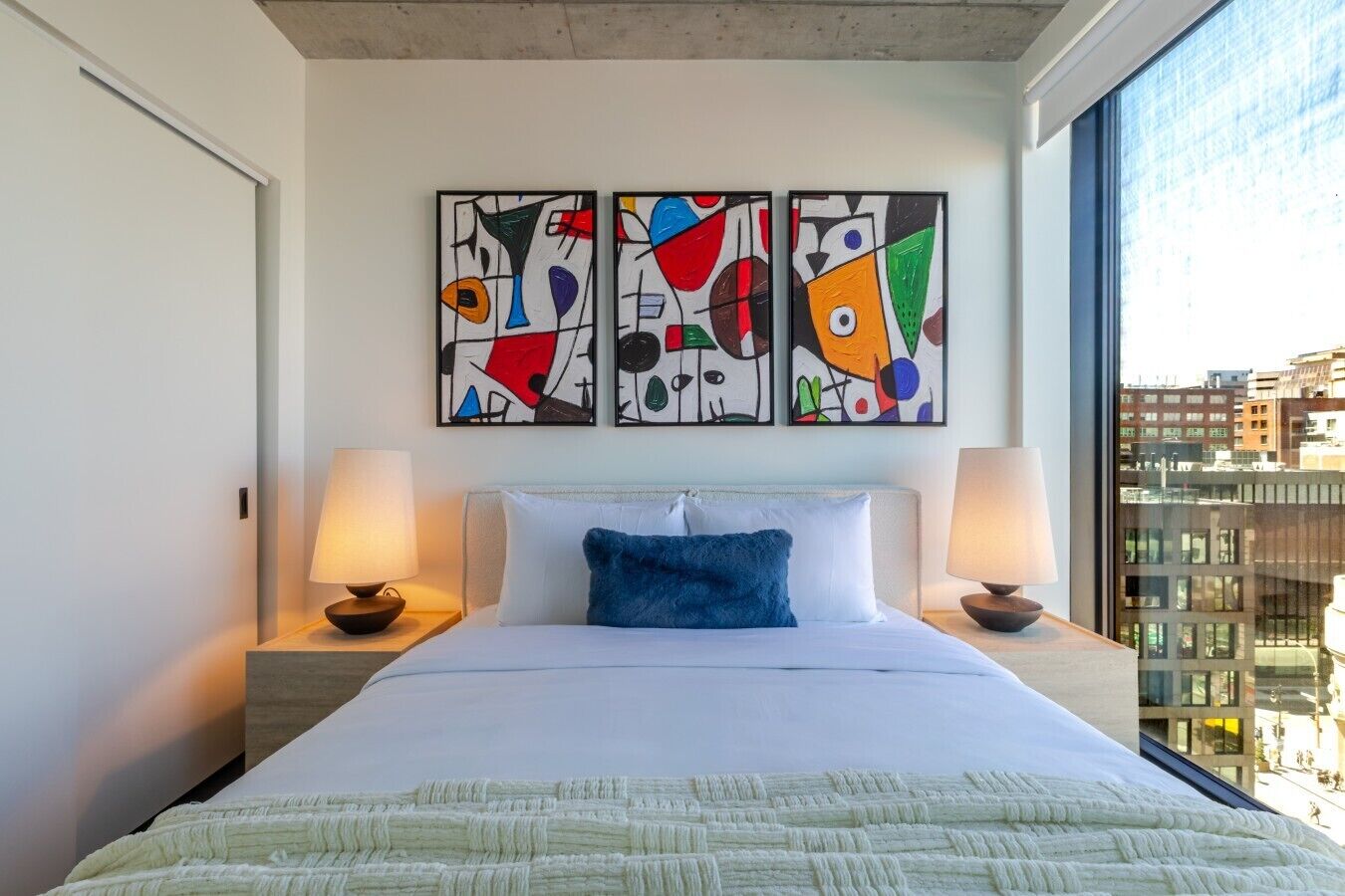 A hotel room with a bed, two bedside lamps, abstract wall art, and a city view.