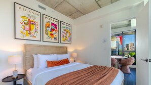 Quarto de hotel com uma cama, uma mesa de cabeceira, uma janela com vista para a cidade e três obras de arte com tema Picasso.