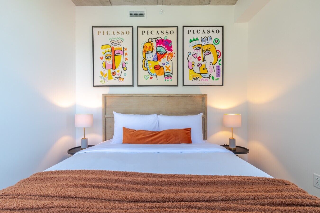 Ein Hotelzimmer mit einem Bett, zwei Nachttischlampen, einer braunen Tagesdecke und drei Kunstwerken von Picasso an der Wand.