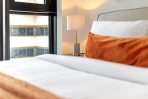 A bed with white bedding, a grey headboard, and an orange pillow. A lamp with a white shade is on a bedside table. - Elegant Downtown Stay | Pools Gym & Rooftop (Montreal)