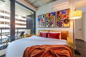 A hotel room with a large bed, a balcony with a view of other buildings, and three abstract paintings on the wall. - Elegant Downtown Stay | Pools Gym & Rooftop (Montreal)