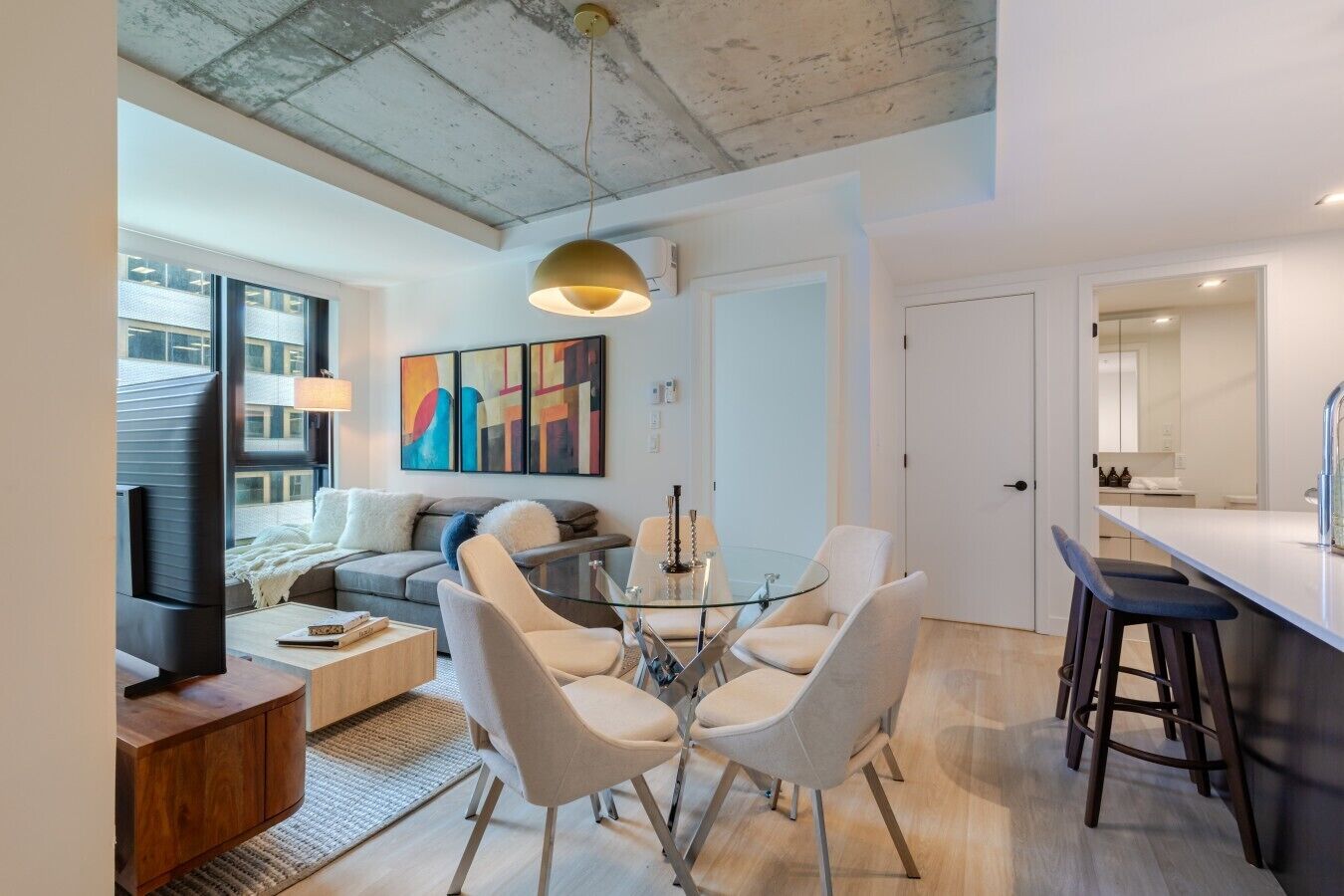 A modern living room with a sofa, coffee table, and TV, a dining area with a glass table and chairs, and a kitchen with a bar.
