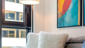 Ein modernes Wohnzimmer mit einem grauen Sofa, weißen, flauschigen Kissen, einer weißen Decke, einem Tisch mit Büchern und einem farbenfrohen, abstrakten Gemälde an der Wand.