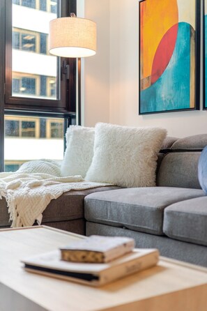 Ein modernes Wohnzimmer mit einem grauen Sofa, weißen, flauschigen Kissen, einer weißen Decke, einem Tisch mit Büchern und einem farbenfrohen, abstrakten Gemälde an der Wand.