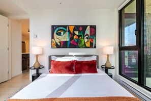 Ein modernes Hotelzimmer mit einem großen Bett, zwei Nachttischen mit Lampen, einem farbenfrohen abstrakten Gemälde über dem Bett und einem großen Fenster mit Blick.
