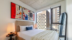 Un hotel moderno con una cama grande, dos lámparas de noche, arte abstracto en la pared y vistas de la ciudad a través de un ventanal.
