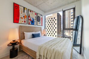 Ein modernes Hotelzimmer mit einem großen Bett, zwei Nachttischlampen, abstrakter Kunst an der Wand und Blick auf die Stadt durch ein großes Fenster.