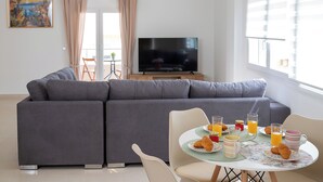 Ein modernes Wohnzimmer mit grauem Sofa, einem Esstisch mit Frühstücksgebäck, einem Flachbildfernseher und einem Gemälde an der Wand.