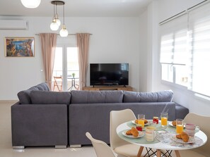 A modern living room with a grey sofa, a dining table with breakfast items, a flat-screen TV, and a painting on the wall. - Maris Apts Kato Gouves (Kato Gouves)
