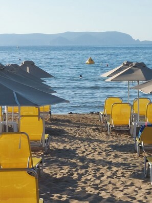 Uma praia com fileiras de cadeiras e guarda-sóis amarelos, areal e mar calmo com montanhas ao longe.