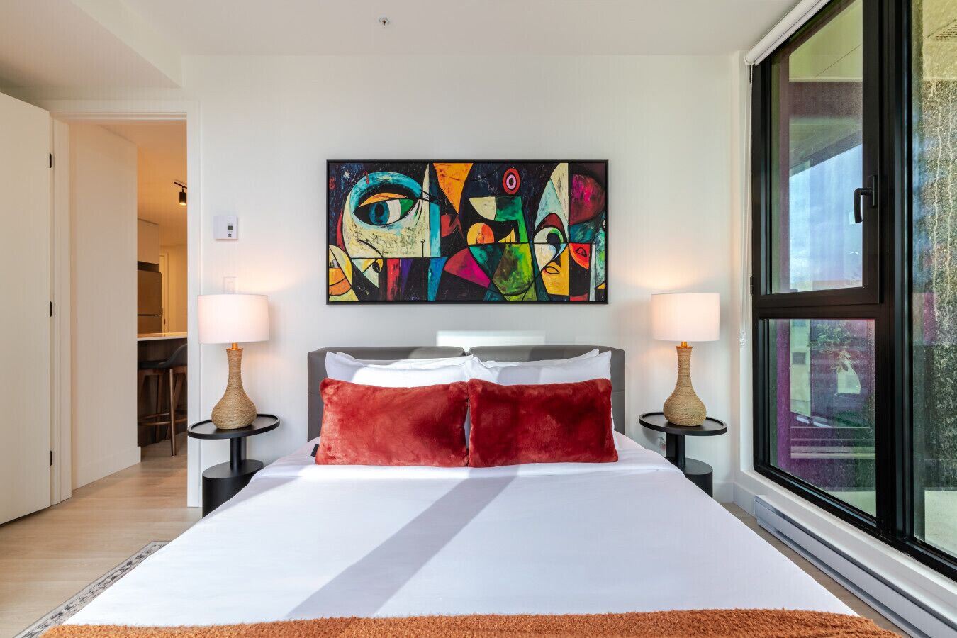 A modern hotel room with a large bed, two bedside tables with lamps, a colorful abstract painting above the bed, and a large window with a view.
