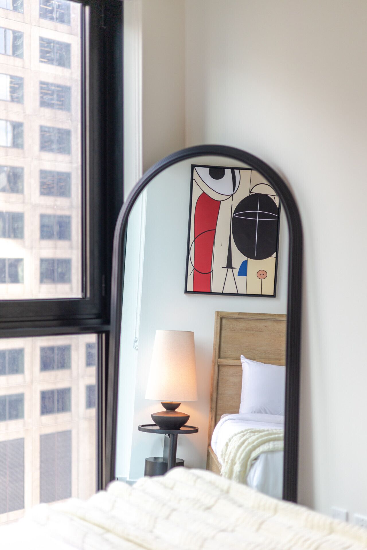 Une chambre d’hôtel avec un lit, une lampe de chevet, un miroir qui reflète un bâtiment et une fenêtre.