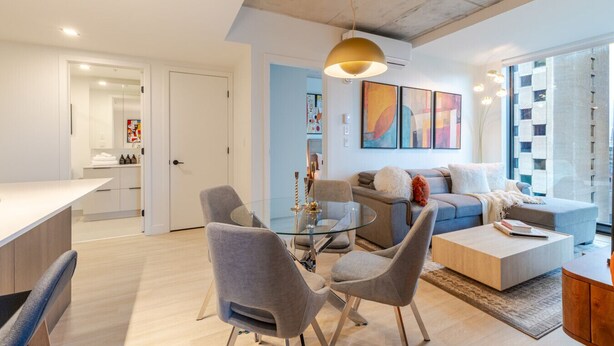 Ett modernt vardagsrum med ett soffbord i glas, grå fåtöljer, en soffa och en matplats med ett vitt bord.