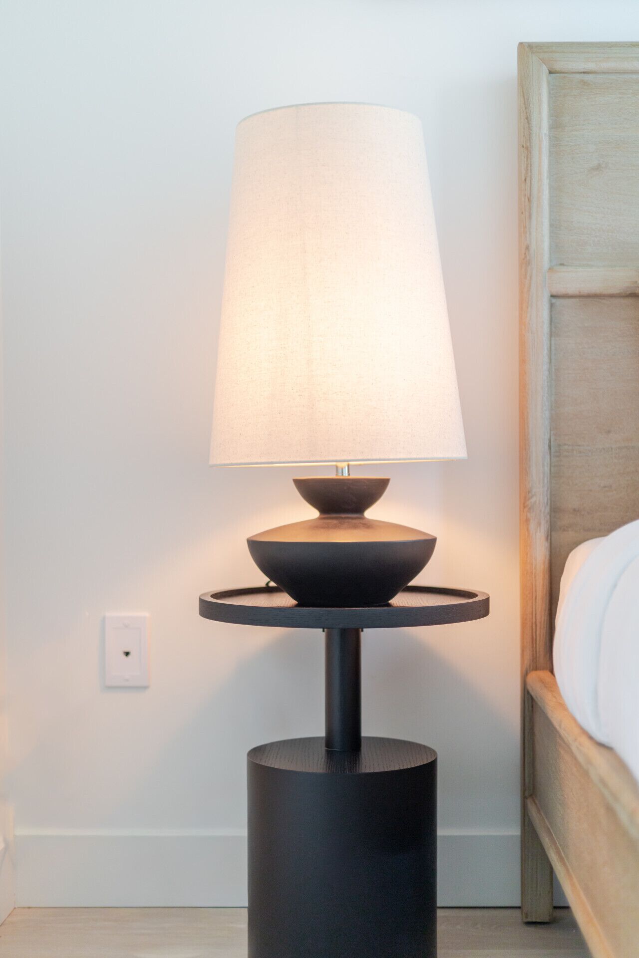A modern bedside table with a lamp, a wooden headboard, and a white wall.