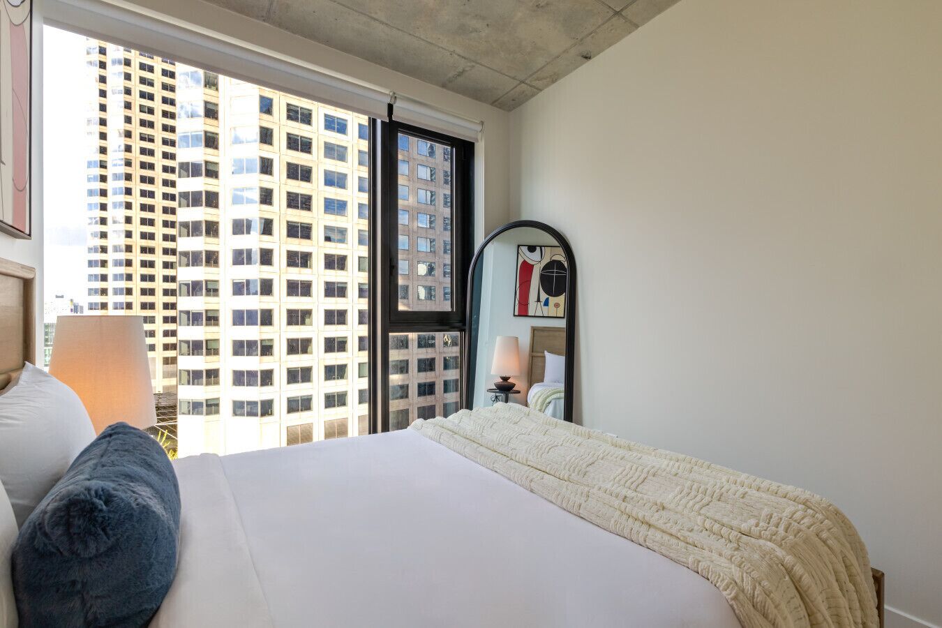 Een hotelkamer met een groot bed, een raam met uitzicht op een hoog gebouw, een nachtkastje met een lamp en een spiegel.