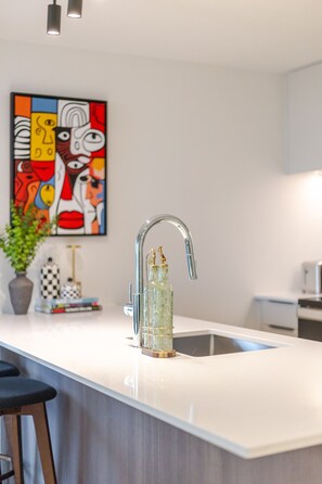 Eine moderne Küche mit weißer Arbeitsplatte, einem Edelstahl-Wasserhahn und einem farbenfrohen, abstrakten Gemälde an der Wand.