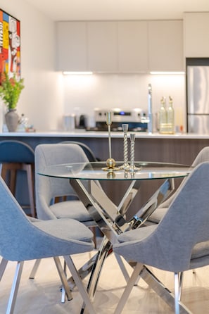A modern kitchen with a glass-top dining table, grey upholstered chairs, stainless steel appliances, and a decorative vase. - Skyline Retreat | Free Parking & Pool (Montréal)