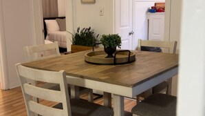 Een eetgedeelte met een houten tafel en witte stoelen, een plant in pot en een witte deur naar een keuken.