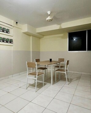 Quarto com mesa de jantar e quatro cadeiras, ventilador de teto, janela e prateleira com objetos.