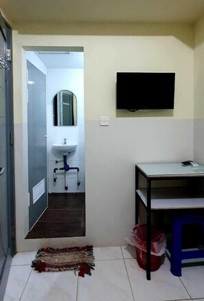 Quarto com pia branca, espelho, televisão presa na parede, mesa pequena e lixeira vermelha.
