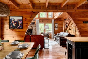 A cozy cabin interior with a dining area, a sofa, a fireplace, and a view of the outdoors. - Luxe Cabin & Guest Cottage w/Views, Firepit & Hot Tub (Sevierville)