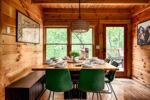 A wooden cabin interior with a dining table set for four, two green chairs, a hanging light fixture, and a large window with a view of trees. - Luxe Cabin & Guest Cottage w/Views, Firepit & Hot Tub (Sevierville)