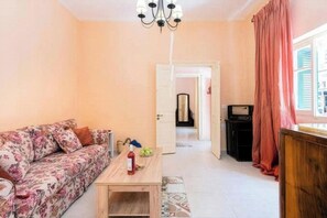 Ein Wohnzimmer mit einem blumigen Sofa, einem Holztisch, einer Chandelieber und Blick durch eine offene Tür.