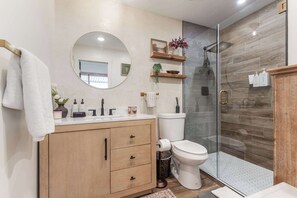 A modern bathroom with a wooden vanity, a round mirror, a toilet, and a glass shower enclosure. - Cowboy Den by the Creek (Swan Valley)