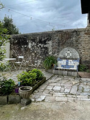 Un mur de pierre, un banc carrelé et un petit espace jardin avec des plantes et une table.