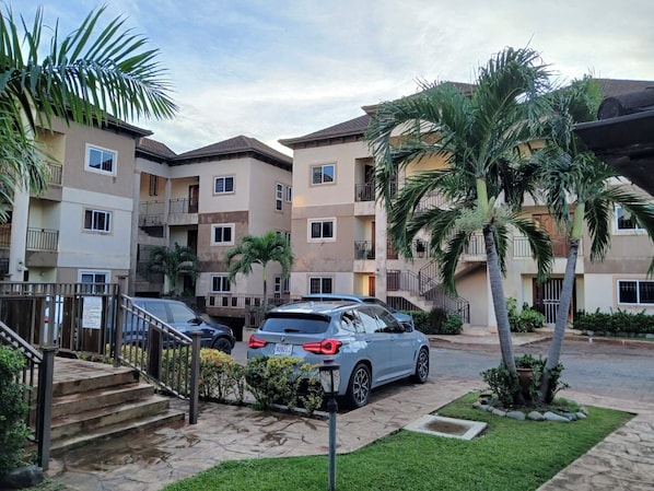 A multi-story residential building with a parking area, palm trees, and a staircase leading to the entrance. - Cozy 2-bedroom apartment in gorgeous Kingston with AC and pool (Kingston)