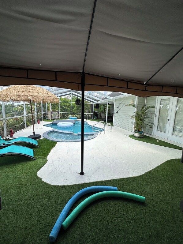 Un espacio exterior cubierto con césped artificial, piscina, sillas de descanso y una sombrilla de paja.