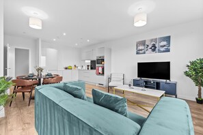 Ein modernes Wohnzimmer mit einem petrolfarbenen Sofa, einem Essbereich, einer Küche und einem Fernseher.