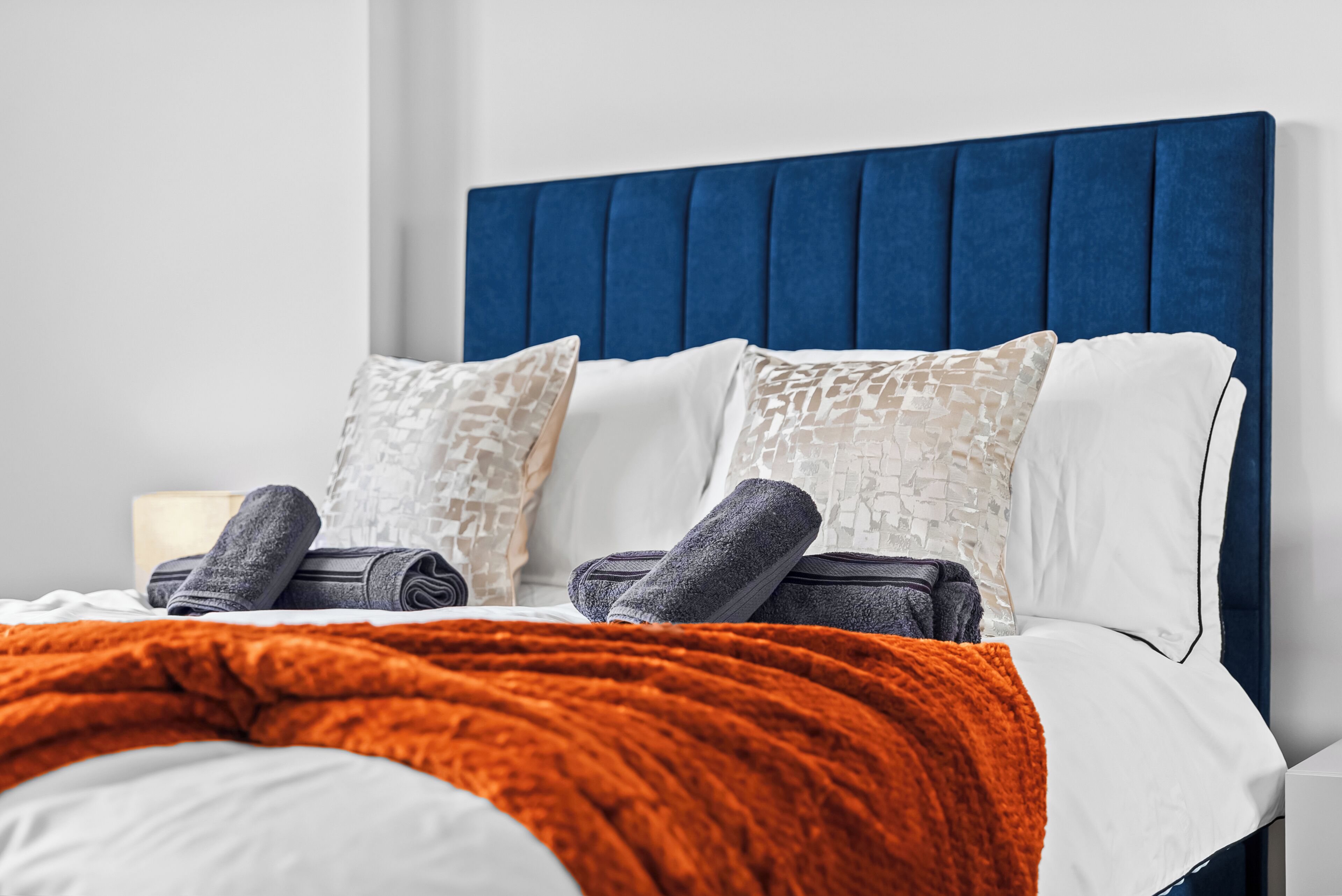 A neatly made bed with a blue headboard, white and patterned pillows, and a folded orange blanket.