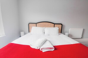 Een net opgemaakt bed met witte lakens en een rode deken, een houten hoofdbord, witte nachtkastjes en een witte radiator.