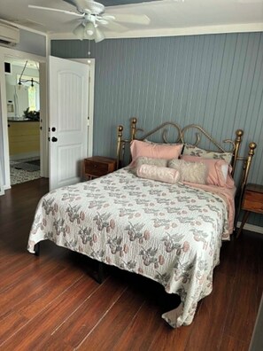 A bedroom with a bed, a nightstand, a wooden floor, a ceiling fan, and a door leading to another room.