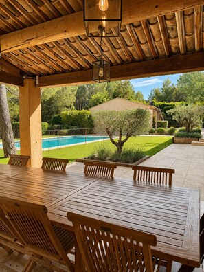 An outdoor patio with a wooden table and chairs, a pool in the background, and a well-maintained garden. - Charming villa in the heart of a golf estate between Luberon and Alpilles (Mallemort)
