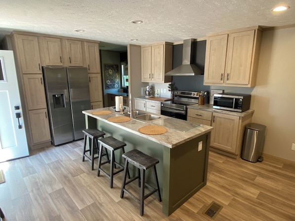 A kitchen with wooden cabinets, a stainless steel refrigerator, an island with stools, and a microwave.