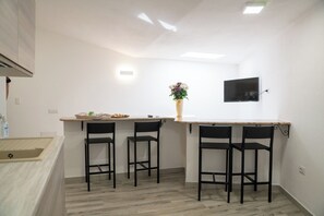 Eine Küche mit einer Holzausziehtafel, zwei schwarzen Barhocker, einer Vase mit Blumen und einem an der Wand befestigten Flachbildfernseher.