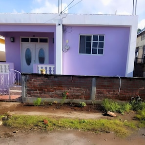 Una casa de dos pisos con fachada morada, una puerta blanca y una ventana pequeña.