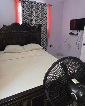 A bedroom with a bed, a wall-mounted TV, and a standing fan. - Jacob’s Urban Haven | 3-Bedroom Home, Goodwill. 4-minute drive to Roseau Central (Roseau)