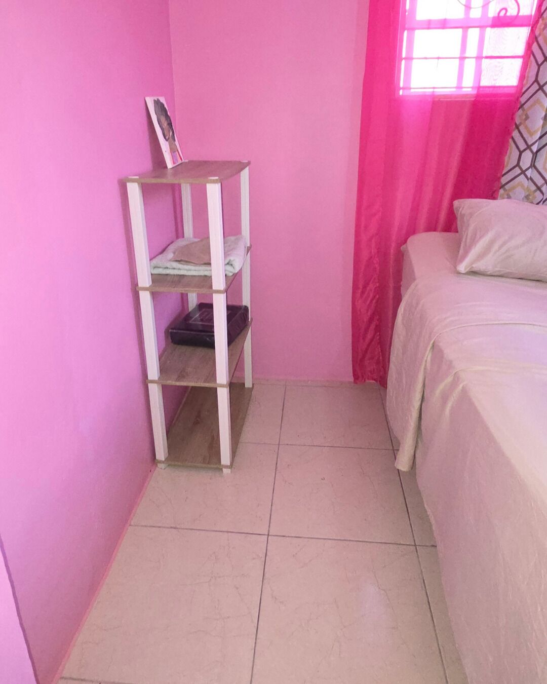 Une chambre avec un lit, une étagère blanche et une fenêtre ornée de rideaux roses.