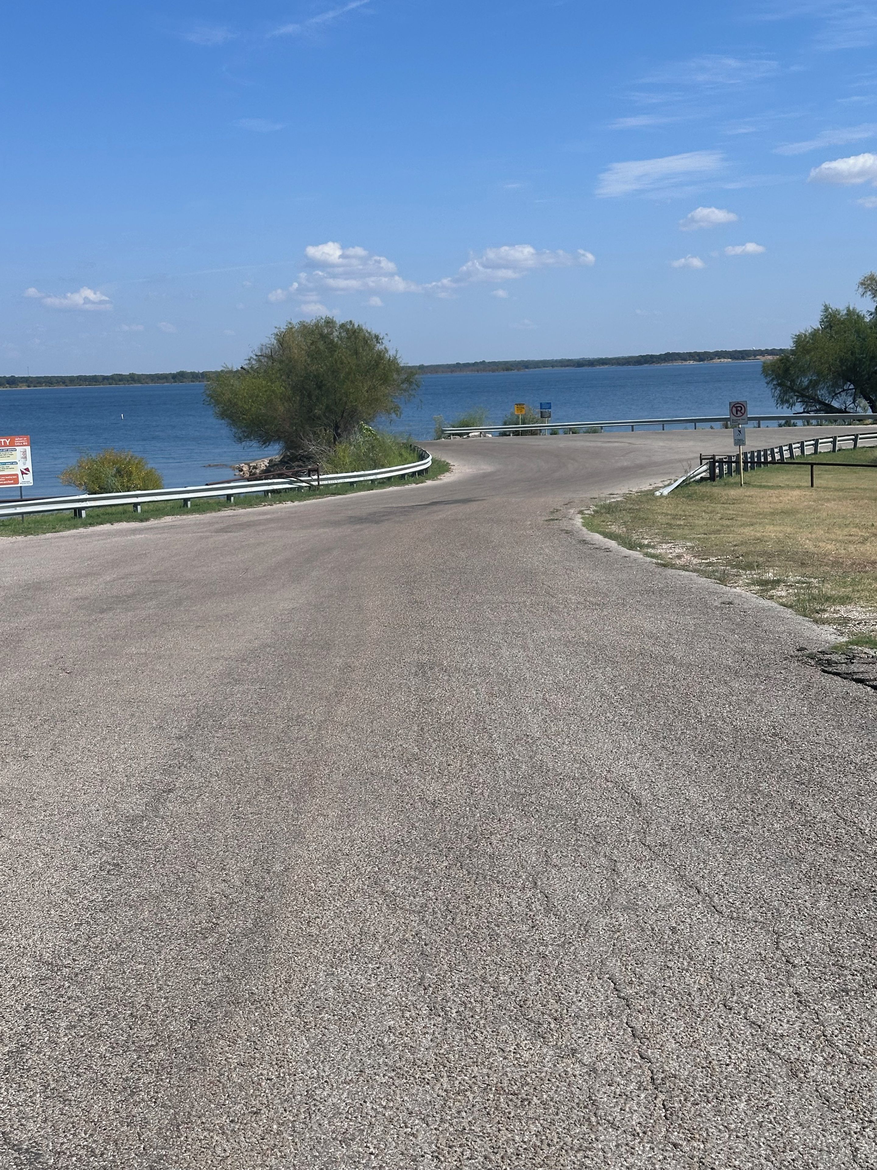 Uma estrada pavimentada serpenteia ao redor de um corpo d'água, com uma placa de proibido estacionar, uma área gramada e um corrimão de metal na beira.