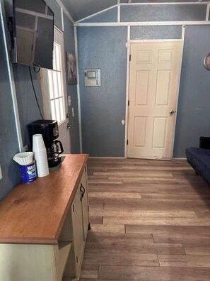 A room with a wooden floor, a white door, a coffee maker, and a can of Maxx House on a counter.