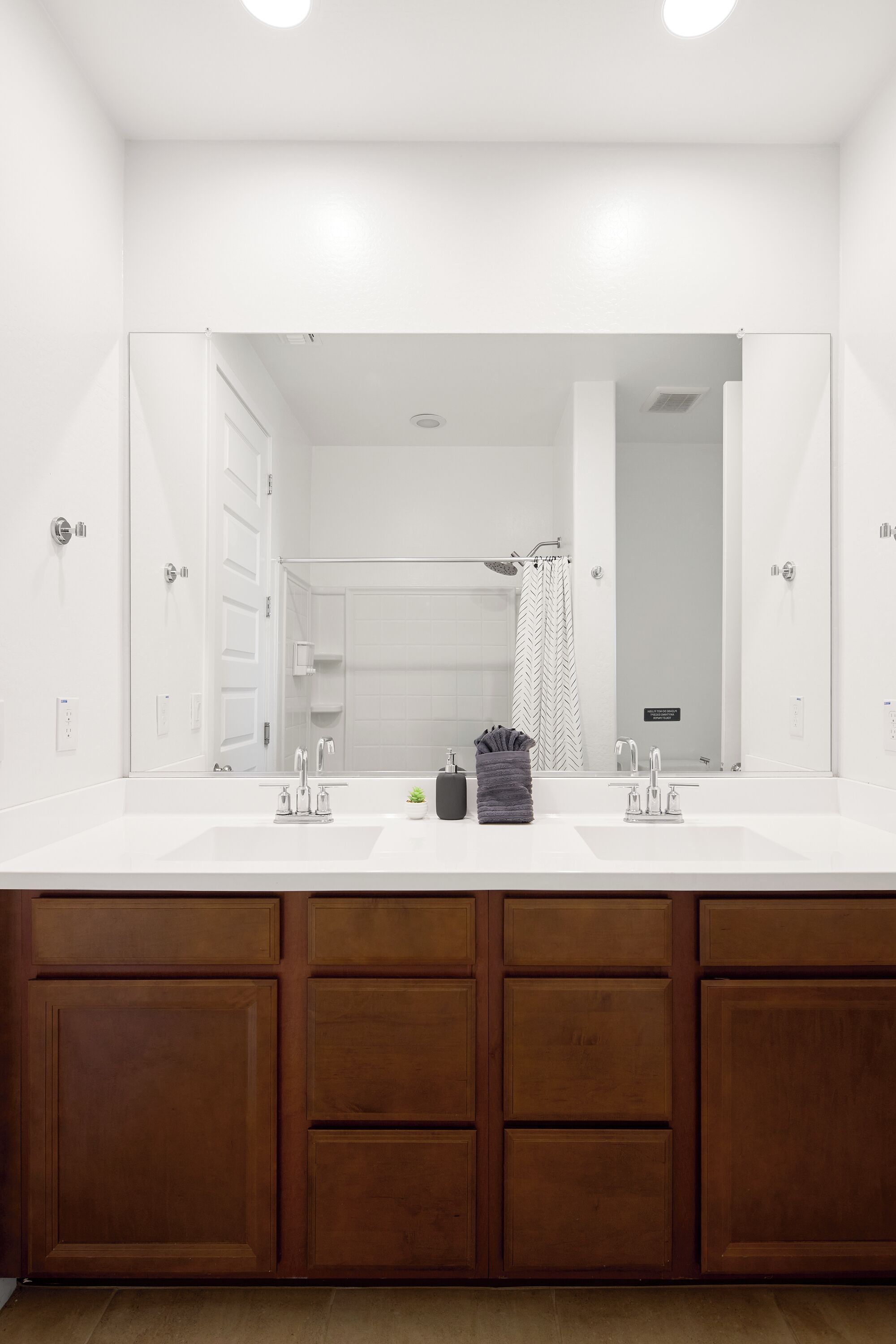 A modern bathroom with a large mirror, two sinks, wooden cabinets, and a shower area.