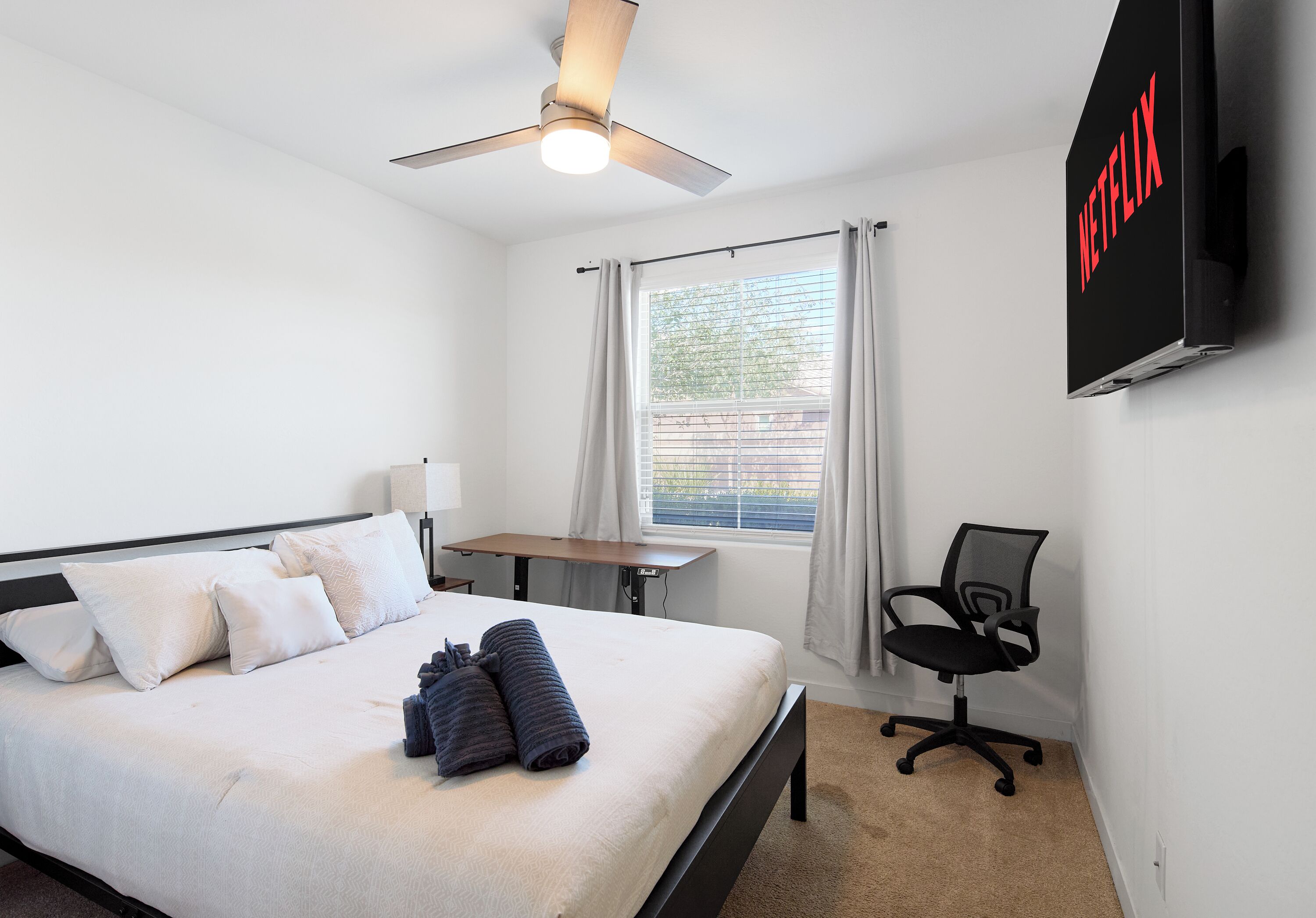 A hotel room with a bed, a desk, a chair, a ceiling fan, a window with curtains, and a TV mounted on the wall.